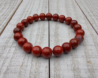 May include: A red jasper beaded bracelet. The bracelet features polished, round beads in varying shades of red and brown. The beads are strung together, forming a circular shape. The bracelet is displayed on a white, weathered wooden surface.