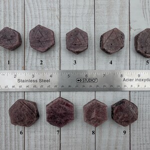 May include: Nine hexagonal, reddish-purple stones with etched designs are arranged on a light wood surface next to a stainless steel ruler. The ruler measures in inches and centimeters. The stones vary in surface texture.