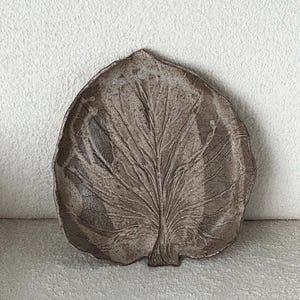 Op de afbeelding: Een bruine keramische schaal in de vorm van een groot blad met een getextureerd oppervlak. De schaal heeft een prominente centrale nerf en kleinere vertakkende nerven, met een onregelmatige, organische rand die een blad nabootst.