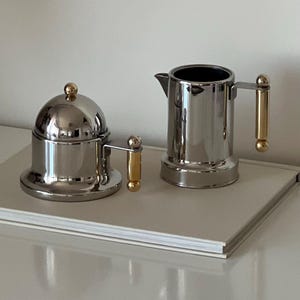 May include: A stainless steel coffee pot and sugar bowl set. The coffee pot has a spout and handle with gold accents. The sugar bowl has a domed lid and a matching handle with gold accents. Both pieces are on a white surface.