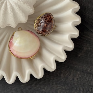Peut inclure: Deux pendentifs en coquillage avec des bords en métal doré. Un coquillage est lisse et rose pâle avec un centre blanc, tandis que l'autre est un cauri brillant tacheté de brun. Les deux ont de petites boucles en métal.