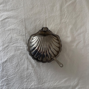 Könnte beinhalten: Eine silberfarbene, muschelförmige Schale mit Griff. Die Schale hat einen gewellten Rand und ein strukturiertes, geriffeltes Inneres. Der Griff ist klein und an der Seite der Muschel befestigt. Die Schale liegt auf einer weißen, zerknitterten Oberfläche.