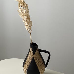 Può includere: Una brocca in ceramica nera opaca con un solo manico e un collo affusolato. Il corpo presenta due pannelli verticali triangolari con un motivo strutturato a linee orizzontali color marrone chiaro. Un singolo stelo fogliato beige chiaro.