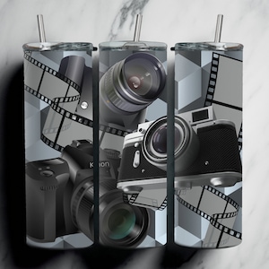 Puede incluir: Tres vasos de acero inoxidable con temática fotográfica. Cada vaso presenta un diseño de cámara diferente, incluyendo una cámara vintage, una cámara moderna y un objetivo de cámara. Los vasos están decorados con un patrón de tira de película en blanco y negro.