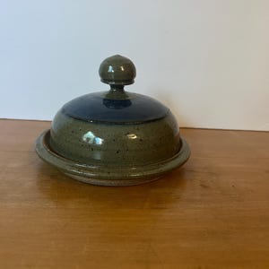 May include: A round ceramic butter dish lid with a speckled olive green base and a smooth dark blue top. The lid features a small, rounded knob handle at the center. The glaze has a glossy finish, and the overall shape is domed with a wide rim.