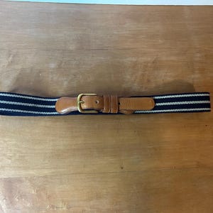 May include: A navy blue and white striped elastic belt with a brown leather strap and a gold-colored buckle. The belt is laid flat on a wooden surface.