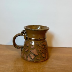 May include: A brown ceramic mug with a unique, abstract design. The mug has a handle that is shaped like a knot.