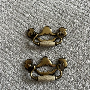 May include: Two antique brass drawer pulls with speckled ceramic handles. The pulls have a decorative design with curved handles and ornate detailing. These hardware pieces are likely for a dresser or cabinet.