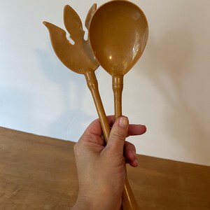 May include: A set of two vintage salad servers in a warm, golden-brown hue. One server is a spoon, the other a fork-like design. The servers are held in a hand against a neutral background, suggesting a kitchen or dining setting.