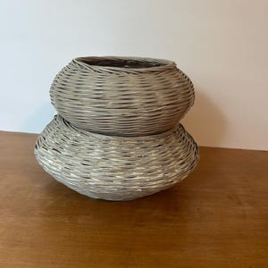 May include: Two woven gray baskets stacked on top of each other. The baskets have a rounded shape and are made of thin, interwoven strips. The baskets are sitting on a wooden surface.