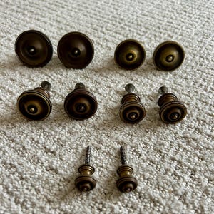 May include: A collection of antique brass cabinet knobs and pulls. The knobs are round with a slightly domed top, while the pulls have a cylindrical design. The hardware is arranged on a neutral, textured surface.