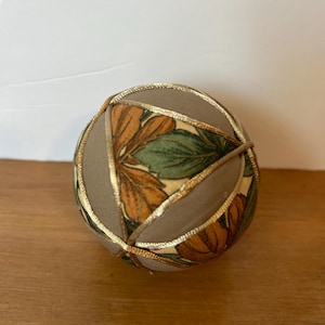 May include: A decorative fabric ball with a geometric design. The ball features segments of tan fabric, accented with gold trim, and panels with a floral pattern in shades of orange, green, and cream. The ball is resting on a wooden surface.