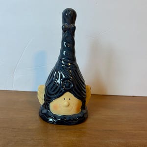 May include: A ceramic figurine with a whimsical design. The figurine features a face with a smiling expression, blonde hair, and small wings. The hair and the top of the figurine are dark blue. The figurine is on a wooden surface.