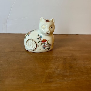 May include: A ceramic cat figurine in a seated position. The off-white cat has floral designs in red, blue, and yellow. A gold swirl design is on the side. The cat has a small brown spot on its head and a smiling expression.