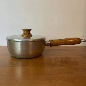 May include: A small, vintage saucepan with a lid. The pan is made of silver-colored metal and has a wooden handle and lid knob. The handle is attached with a metal ring.