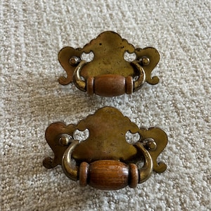 May include: Two antique brass drawer pulls with wooden handles. The brass hardware has a decorative, ornate design with a curved handle. The wooden handle is cylindrical and has a warm, brown color. These pulls are likely for furniture.