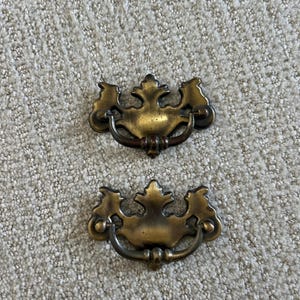 May include: Two matching brass drawer pulls featuring a decorative, ornate backplate with a central fleur-de-lis inspired motif and a curved, swinging bail handle. The metal has a darkened, aged patina with visible highlights on the edges.