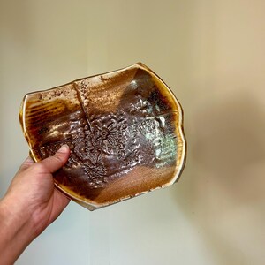 May include: A handmade, ceramic serving dish with a unique, organic shape. The dish features a rich, brown glaze with lighter brown and cream accents. The surface is textured with a floral pattern, adding visual interest to the piece.