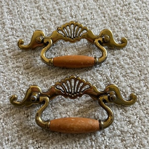 May include: Two vintage-style brass drawer pulls with wooden handles. The brass hardware features a decorative fan-shaped design and ornate scrollwork. The wooden handles are a light brown color. These pulls are likely for a cabinet or dresser.