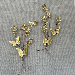 May include: Two decorative metal wall art pieces featuring gold-colored butterflies and flowers. The butterflies have detailed wing patterns and are attached to thin, brown stems with gold flowers. The pieces are mounted on a textured, light gray background.