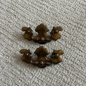 May include: Two antique brass drawer pulls with a decorative design. Each pull has a central, ornate handle with two smaller, curved handles on either side. The pulls are set against a neutral, textured background.