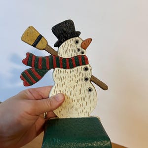 May include: A wooden snowman Christmas stocking holder. The snowman has a black top hat, a carrot nose, and a red and green striped scarf. It holds a yellow broom and has a green base.