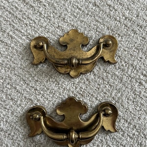 May include: Two antique brass drawer pulls. Each pull features a curved handle with decorative floral-like accents on each end. The pulls are a warm, aged brass color, suggesting vintage or retro design.