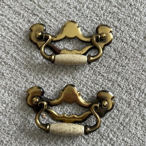 May include: Two antique brass drawer pulls with cream-colored speckled ceramic handles. The pulls have a decorative, ornate design with curved handles and a vintage aesthetic. Perfect for adding a touch of elegance to furniture.