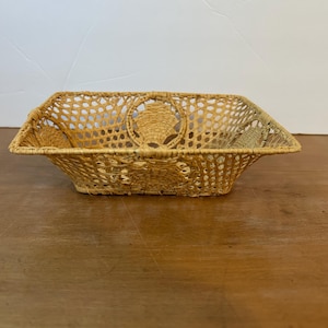 May include: A rectangular woven basket with a light tan color. The basket has a decorative open weave design with a scalloped edge. The basket is sitting on a wooden surface.