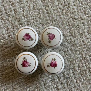 May include: Four white ceramic cabinet knobs with a gold rim and a pink rose design. The knobs are round and have a classic, floral aesthetic, suitable for furniture restoration or adding a vintage touch to home decor.