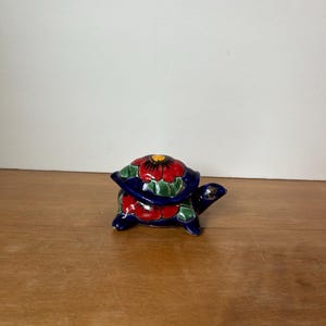 May include: A small, hand-painted ceramic turtle figurine with a blue shell, red and green flowers, and a yellow center. The turtle is sitting on a wooden surface.