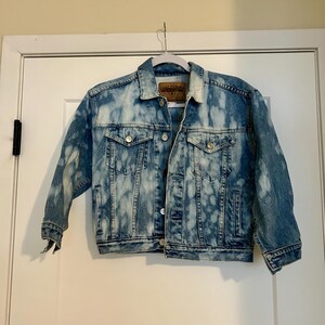 May include: A light blue denim jacket with a bleached effect. The jacket has a button closure and two pockets on the front.