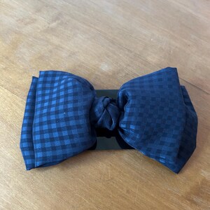 May include: A navy blue bow tie with a checkered pattern. The bow tie is made of fabric and is attached to a black band. The bow tie is laying on a wooden surface.