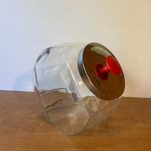 May include: A clear glass jar with a wide, rounded body and a metal lid. The lid has a red knob. The jar is empty and sits on a wooden surface. The jar is designed for storage.
