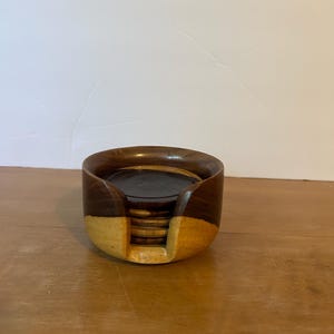 May include: A wooden coaster set with a holder. The holder is a rounded shape, with a dark brown top and a lighter, natural wood color on the bottom. The coasters are stacked inside the holder, and are a similar color to the top of the holder.