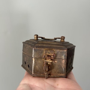 May include: A small, antique brass box with an octagonal shape. The box features a hinged lid with a latch closure and decorative accents. The brass has a tarnished, aged appearance, suggesting its vintage. The box is held in a hand.