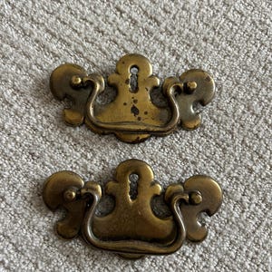 May include: Two antique brass drawer pulls with a keyhole design. Each pull features a curved handle and decorative side elements. The brass has a weathered appearance, suggesting age and use. These pulls are likely for a vintage cabinet or drawer.