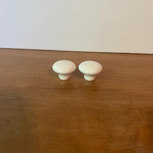 May include: Two white ceramic cabinet knobs with a rounded top and a short, cylindrical base. The knobs are set against a wooden surface and a white background. These knobs are suitable for furniture restoration or DIY projects.