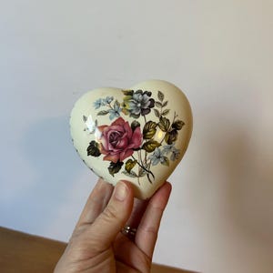 May include: Heart-shaped trinket box with a floral design. The box is cream-colored and features a bouquet of roses in shades of pink and red, with blue and purple accent flowers and green leaves. The box is held in a hand.