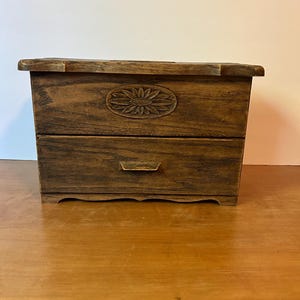 May include: A rectangular wooden box with a dark brown finish, featuring a single drawer with a horizontal handle and a carved oval floral motif centered on the upper front panel. The box has a flat top and a scalloped base design.
