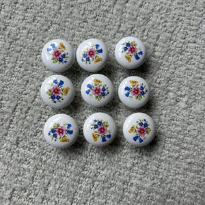 May include: A set of nine round white ceramic knobs, each featuring a central floral pattern with pink, blue, and yellow flowers and green leaves. The knobs are arranged in a three-by-three grid, showcasing a smooth, glossy finish on each piece.