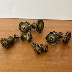 May include: A collection of five antique brass cabinet knobs with a circular, tiered design and a dark, weathered finish. Each knob features a central raised point and a ridged outer edge, accompanied by threaded metal screws for installation.