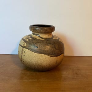 May include: A round ceramic vase with a short, thick neck and a wide opening. The surface features a speckled beige base color with dark brown, abstract, flowing glaze patterns that wrap around the body and a series of small dots on one side.