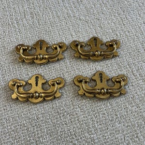 May include: A set of four brass drawer pulls, each featuring a decorative backplate with a central keyhole cutout and a curved, swinging handle. The metal hardware has a polished finish with subtle darkened accents in the recessed areas.