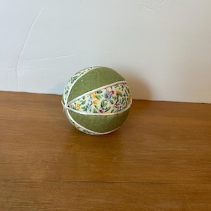 May include: A handmade fabric ball with a floral pattern and green sections. The ball is round and has a white ribbon detail. The ball is sitting on a wooden surface.