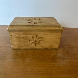 May include: A small, rectangular wooden box with a carved floral design on the lid and front. The box is made of light-colored wood and has a simple, classic design. The box is sitting on a wooden surface.
