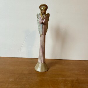 May include: A slender angel figurine with a gold halo and wings, dressed in a pink and light blue gown. The angel is standing on a beige base. The figurine is on a wooden surface.