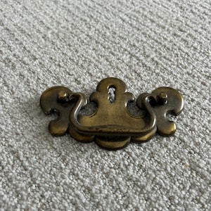 May include: A vintage-style brass drawer pull with a decorative design. The pull features a central handle with a keyhole and two rounded end pieces. The hardware is likely for a cabinet or dresser.