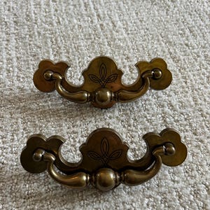 May include: Two antique brass drawer pulls. Each pull features a decorative floral design on the center plate and curved handles. The pulls are set against a neutral, textured background.