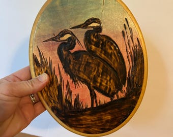 May include: Oval wooden plaque featuring a pyrography artwork of two herons in a marsh setting. The birds are depicted in shades of brown, with a backdrop of pink and blue hues. The plaque has a natural wood border.